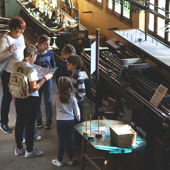 Groupe d'enfants avec leurs parents dans un atelier de filature de laine, découvrant les anciennes machines de la Filature de Laine de Belvès.